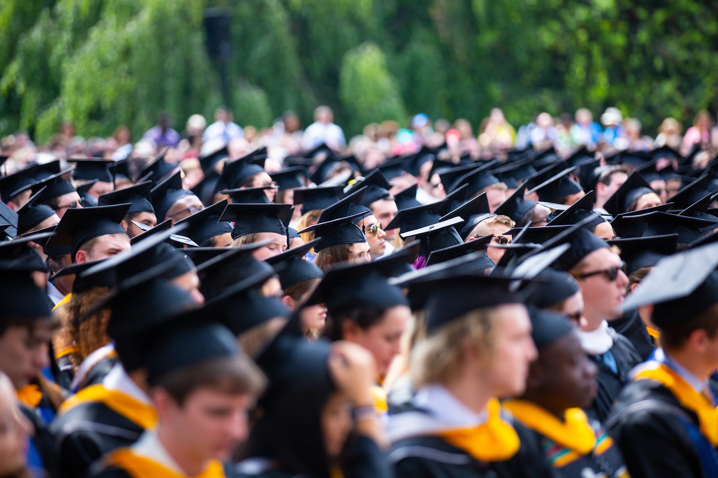 A large group of Georgetown graduates wearing caps and gowns, seated outdoors during a graduation ceremony, with trees and a crowd of people in the background.