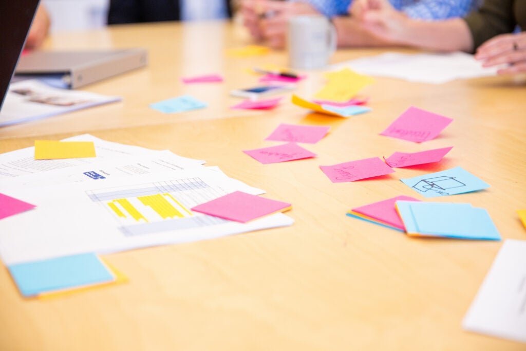 Table filled with colorful sticky notes, papers, and binders. People sitting around the table in the background.